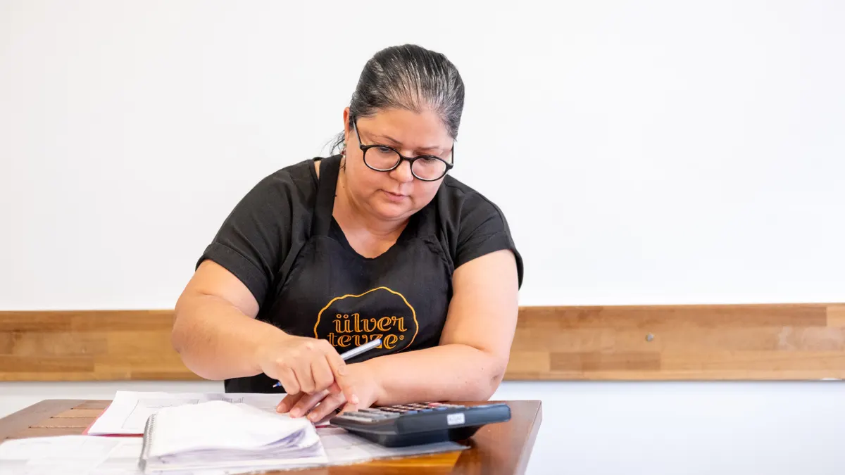 Femme d'âge moyen utilisant une calculatrice pour des tâches financières à un bureau en bois, à l'intérieur.