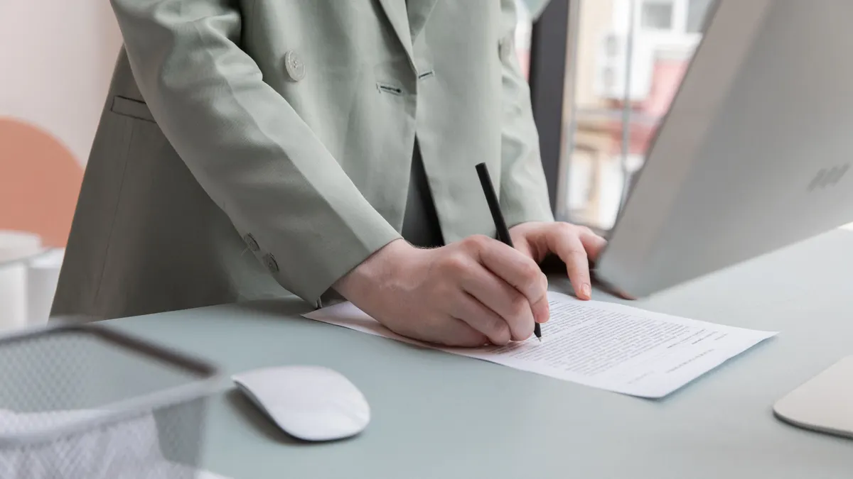 Personne non identifiable debout à une table avec un ordinateur, prenant des notes sur un document au bureau